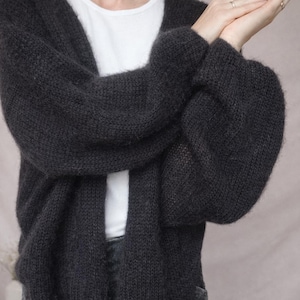 May include: Dark gray open-front cardigan with long sleeves. The sweater is made of a chunky knit material and is worn over a white t-shirt. The cardigan has a relaxed fit and is suitable for layering. The sleeves are voluminous.