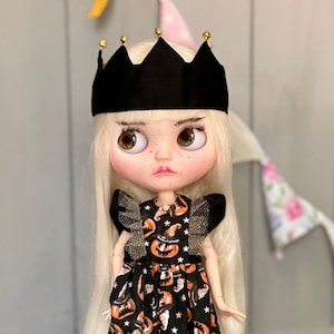 Könnte beinhalten: Eine Puppe mit blonden Haaren, die eine schwarze Krone, ein schwarzes und orangefarbenes Kleid mit Kürbis- und Hexenhutmuster und türkisfarbene Schuhe trägt.
