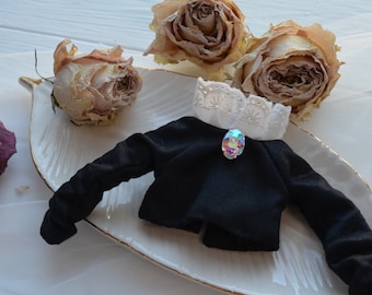 May include: A black cropped jacket with long sleeves and a white lace collar. A clear iridescent jewel is pinned to the front of the jacket.