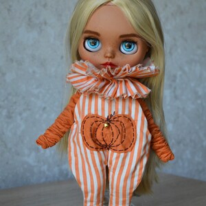 May include: A doll with blonde hair wearing an orange and white striped jumpsuit with a pumpkin design. The doll has large blue eyes and a painted face.