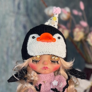 Può includere: Una bambola con i capelli biondi che indossa un cappello da pinguino bianco e nero con un becco arancione. La bambola indossa un maglione rosa e una gonna di tulle rosa con fiori bianchi.