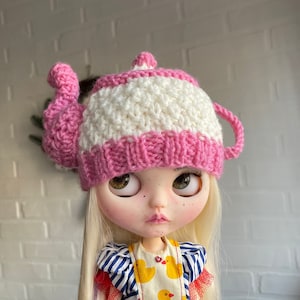 Puede incluir: Una muñeca con un gorro de tetera tejido en rosa y blanco. El gorro tiene una parte superior blanca y una parte inferior rosa con un asa tejida blanca.