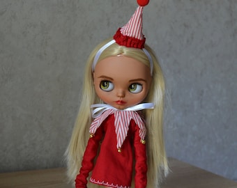 May include: A doll with blonde hair wearing a red and white striped dress with a white collar and a red and white striped hat with a red pom-pom on top. The doll is wearing purple shoes with white soles.