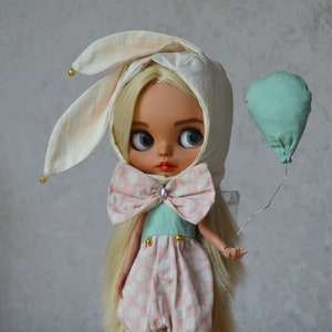 May include: A doll with long blonde hair wearing a cream-coloured hood with long rabbit ears and small gold beads. The doll wears a light green bodice and pink polka-dot trousers with a large matching bow at the neck and holds a mint green balloon.