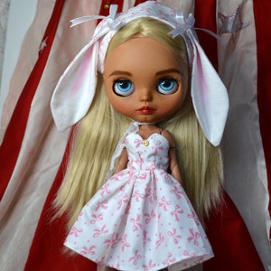 May include: A Blythe doll with blonde hair and blue eyes, wearing a white dress with pink bows. The doll has a white headband with attached bunny ears. The background features red and white striped fabric.