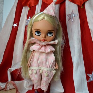 Op de afbeelding: Een blonde pop met een roze en witte sterrenprint jumpsuit met een witte ruche kraag en een witte hoed. De pop staat voor een rood-wit gestreepte stof met zilveren sterren.