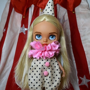 Könnte beinhalten: Eine blonde Puppe, die einen schwarz-weißen Polka-Dot-Overall mit einem rosa Rüschenkragen und einer schwarz-weißen Polka-Dot-Mütze trägt. Die Puppe hat blaue Augen und steht vor einem rot-weiß gestreiften Zirkuszelt.