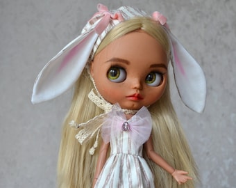 Könnte beinhalten: Eine Puppe mit langen blonden Haaren, die ein weißes Kleid mit rosa Streifen, ein weißes Stirnband mit rosa Schleifen und weiße Hasenohren trägt.
