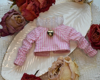 Op de afbeelding: Een roze en wit gestreept poppenoverhemd met een gouden hartbedel op een wit bord omringd door gedroogde rozen.
