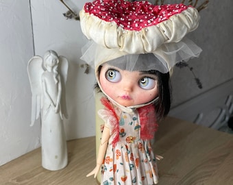 May include: A doll with black hair wearing a white dress with a mushroom print and a red and white mushroom hat. The doll has blue eyes and is wearing purple shoes.
