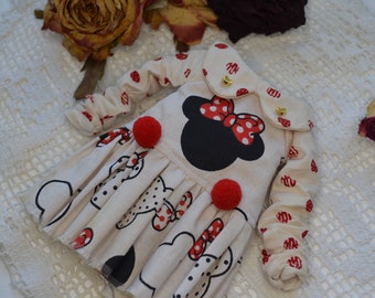 May include: A white dress with red polka dots and a black and red Minnie Mouse design. The dress has a white collar with gold buttons and red pom-poms on the sleeves.