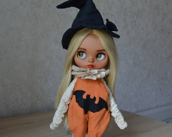 May include: A doll wearing a witch's hat and a pumpkin-themed outfit with a bat design. The doll has long blonde hair and large green eyes.