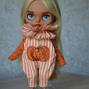 May include: A doll with long blonde hair wearing an orange and white striped jumpsuit with a pumpkin design. The doll has blue eyes and a freckled face.