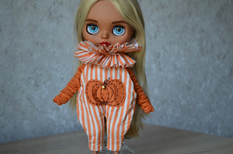 May include: A doll with long blonde hair wearing an orange and white striped jumpsuit with a pumpkin design. The doll has blue eyes and a freckled face.