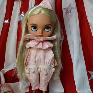 Op de afbeelding: Een blonde pop met een roze en witte sterrenprint jumpsuit met een gerimpelde kraag en een witte hoed. De pop staat voor een rood-wit gestreepte stof met witte sterren.