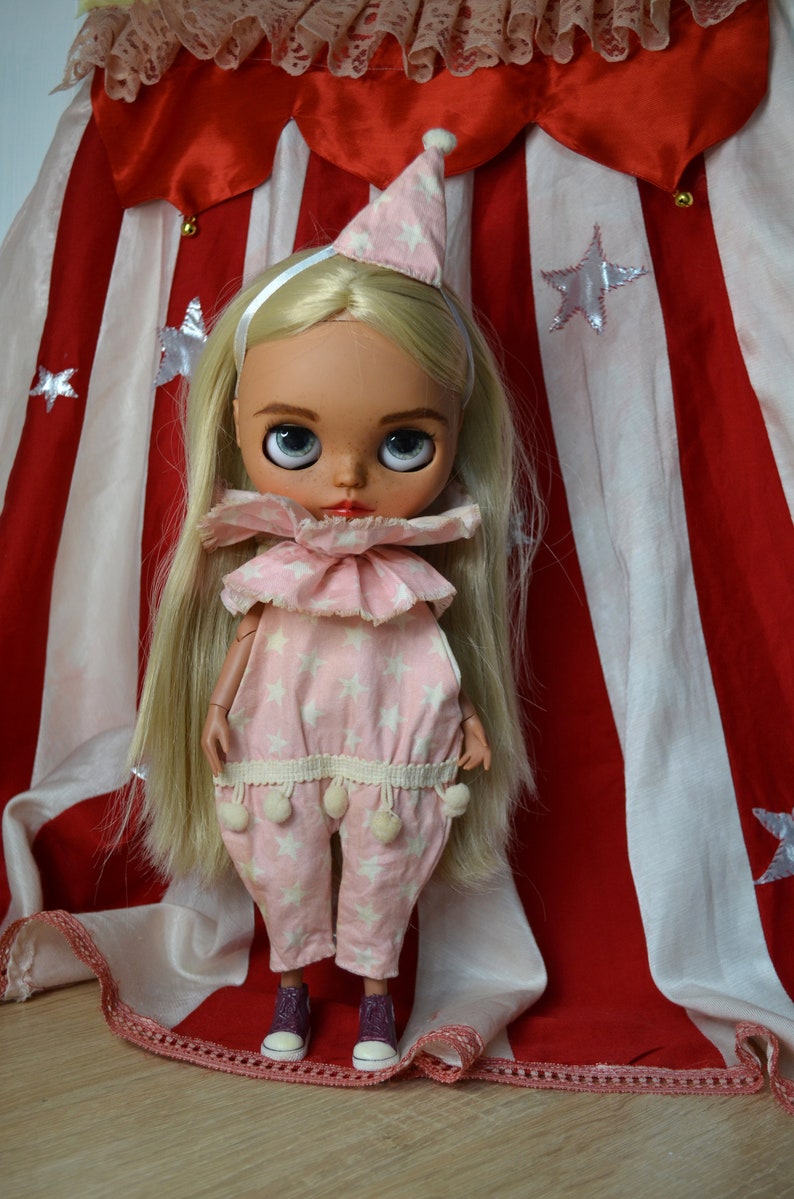 Op de afbeelding: Een blonde pop met een roze en witte sterrenprint jumpsuit met een gerimpelde kraag en een witte hoed. De pop staat voor een rood-wit gestreepte stof met witte sterren.