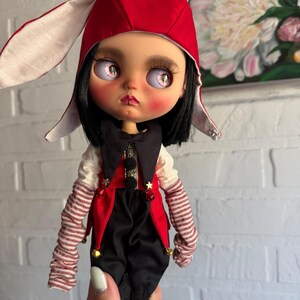 Pu&ograve; includere: Una bambola con capelli neri e occhi rosa pallido che indossa un cappuccio da coniglio rosso e bianco, un gilet nero e rosso con piccoli ciondoli a forma di stella dorata, una camicia bianca a maniche lunghe, scaldabraccia a righe e pantaloni neri.