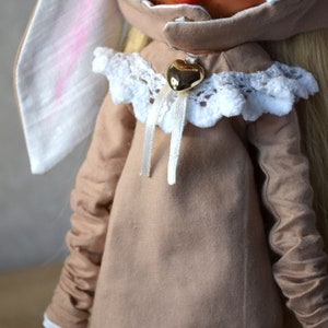 Puede incluir: Una mu&ntilde;eca marr&oacute;n que lleva un vestido marr&oacute;n con ribete de encaje blanco y un dije de coraz&oacute;n dorado. La mu&ntilde;eca tiene el pelo largo y rubio y orejas de conejo blancas.