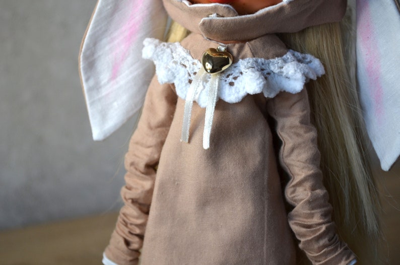 Puede incluir: Una mu&ntilde;eca marr&oacute;n que lleva un vestido marr&oacute;n con ribete de encaje blanco y un dije de coraz&oacute;n dorado. La mu&ntilde;eca tiene el pelo largo y rubio y orejas de conejo blancas.