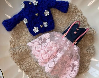 Könnte beinhalten: Ein miniaturer blauer Strickpullover mit weißen Blumen- und goldenen Herzverzierungen, neben einem rosa Spitzenkleid mit marineblauem Oberteil und rosa Trägern. Die Artikel werden auf einem dekorativen Teller präsentiert.