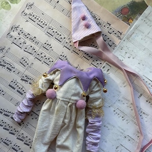 Può includere: Un costume da giullare viola e bianco con un cappello, campanelli e pon pon. Il costume è fatto di tessuto e si trova su un foglio di musica.
