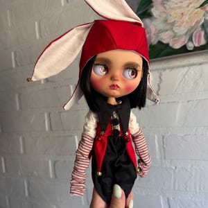 Pu&ograve; includere: Una bambola con capelli neri e occhi rosa chiaro indossa un cappello da giullare rosso e bianco con piccoli campanelli dorati. La bambola ha un gilet nero e rosso sopra una camicia bianca con maniche a righe rosse e bianche, pantaloni neri.