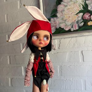 Pu&ograve; includere: Una bambola con capelli neri e grandi occhi chiari indossa un cappello rosso e bianco con orecchie da coniglio e piccoli campanelli. La bambola indossa un gilet nero e rosso sopra una camicia bianca con maniche a righe rosse e bianche e stelle.