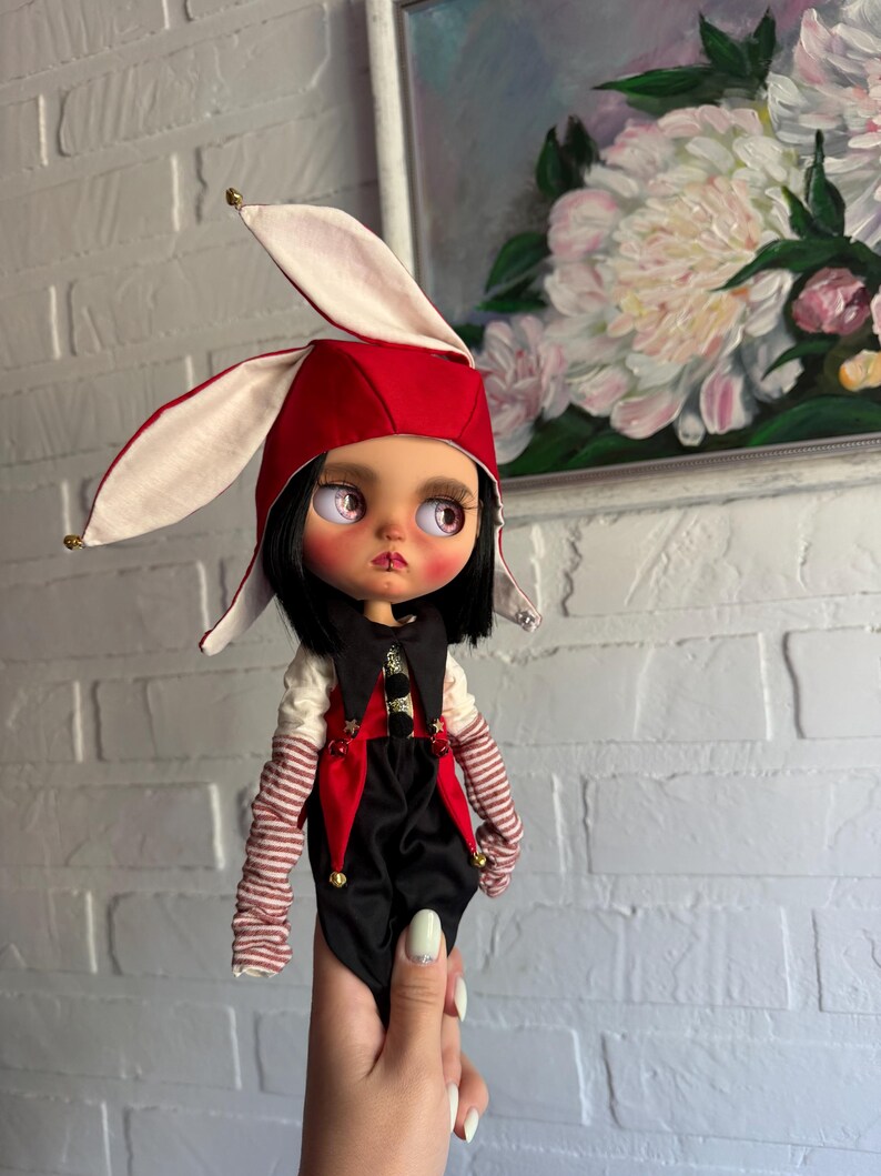 Pu&ograve; includere: Una bambola con capelli neri e grandi occhi chiari indossa un cappello rosso e bianco con orecchie da coniglio e piccoli campanelli. La bambola indossa un gilet nero e rosso sopra una camicia bianca con maniche a righe rosse e bianche e stelle.