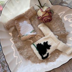 Puede incluir: Dos jerséis de punto en miniatura, uno beige con la silueta de un gato blanco y otro blanco con la silueta de un gato negro, ambos con una fina línea horizontal verde cerca del dobladillo inferior, expuestos junto a una rosa seca.