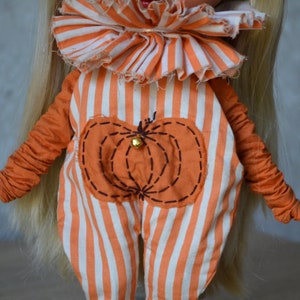 May include: A doll wearing an orange and white striped jumpsuit with a pumpkin design on the front. The jumpsuit has a ruffled collar and the pumpkin has a small bell in the centre.