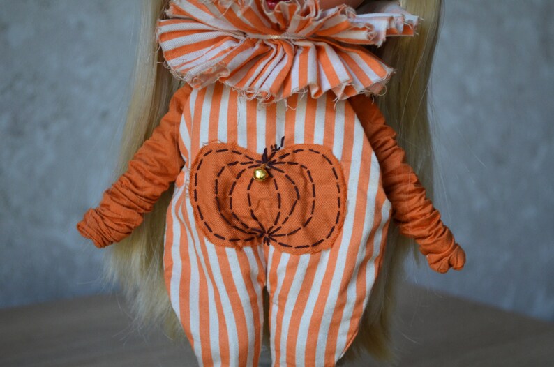 May include: A doll wearing an orange and white striped jumpsuit with a pumpkin design on the front. The jumpsuit has a ruffled collar and the pumpkin has a small bell in the centre.