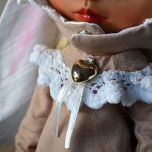 Puede incluir: Una mu&ntilde;eca con un vestido marr&oacute;n con ribete de encaje blanco y un colgante de coraz&oacute;n dorado. La mu&ntilde;eca tiene el pelo rubio y lleva una diadema de orejas de conejo blancas.