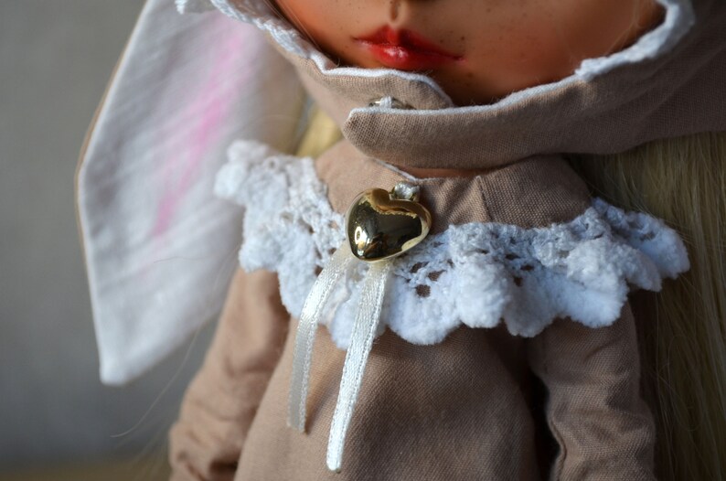 Puede incluir: Una mu&ntilde;eca con un vestido marr&oacute;n con ribete de encaje blanco y un colgante de coraz&oacute;n dorado. La mu&ntilde;eca tiene el pelo rubio y lleva una diadema de orejas de conejo blancas.