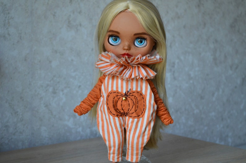 May include: A doll with blonde hair wearing an orange and white striped jumpsuit with a pumpkin design. The doll has large blue eyes and a painted face.