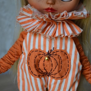 May include: A doll wearing an orange and white striped jumpsuit with a pumpkin patch design. The jumpsuit has a ruffled collar and the doll has blonde hair.