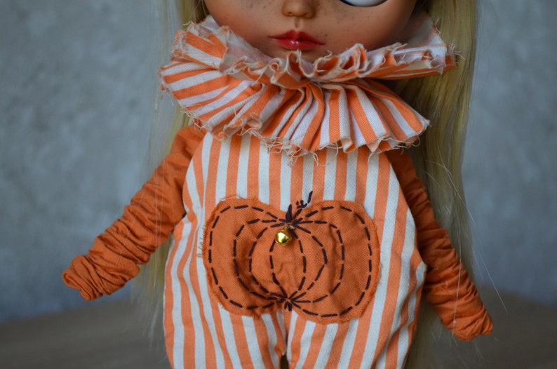 May include: A doll wearing an orange and white striped jumpsuit with a pumpkin patch design. The jumpsuit has a ruffled collar and the doll has blonde hair.