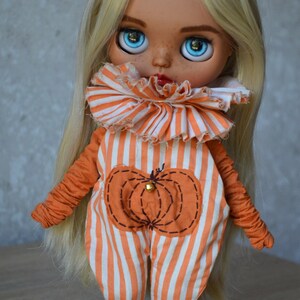 May include: A blonde doll wearing a white and orange striped jumpsuit with a pumpkin design. The doll has blue eyes and freckles.