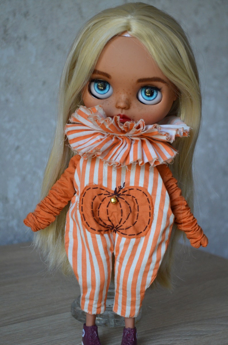 May include: A blonde doll wearing a white and orange striped jumpsuit with a pumpkin design. The doll has blue eyes and freckles.
