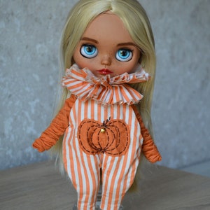 May include: A blonde doll wearing an orange and white striped jumpsuit with a pumpkin design. The doll has blue eyes and is wearing maroon trainers.
