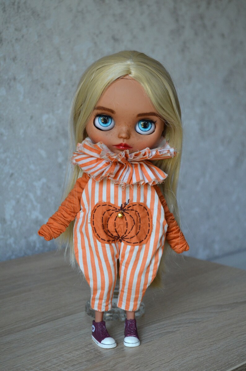 May include: A blonde doll wearing an orange and white striped jumpsuit with a pumpkin design. The doll has blue eyes and is wearing maroon trainers.