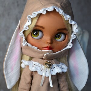 Puede incluir: Una mu&ntilde;eca con cabello rubio que lleva un atuendo marr&oacute;n con capucha, ribete de encaje blanco y orejas de conejo. La mu&ntilde;eca tiene ojos verdes y pecas.