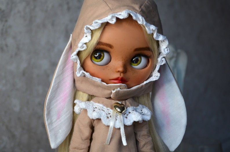Puede incluir: Una mu&ntilde;eca con cabello rubio que lleva un atuendo marr&oacute;n con capucha, ribete de encaje blanco y orejas de conejo. La mu&ntilde;eca tiene ojos verdes y pecas.