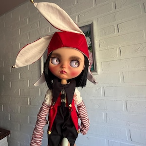 Pu&ograve; includere: Una bambola con capelli neri e grandi occhi rosa indossa un cappello da giullare rosso e bianco con lunghe orecchie. La bambola veste un gilet nero e rosso con piccoli campanelli dorati, una camicia bianca con maniche a righe rosse e bianche.