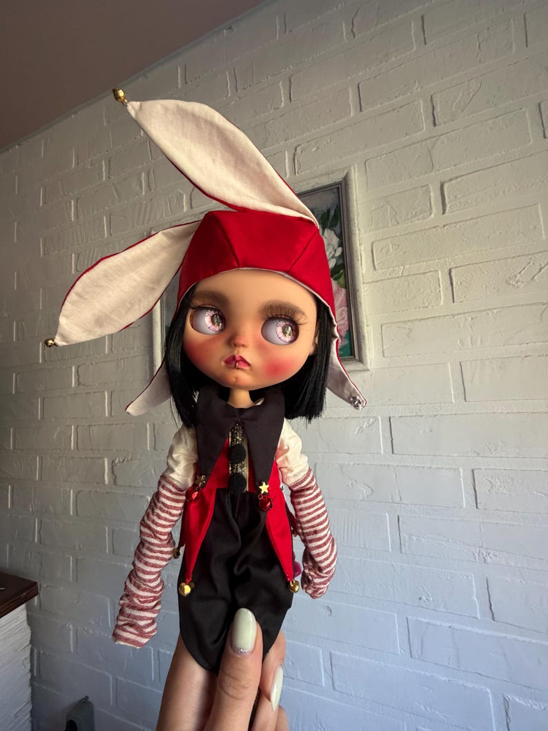Pu&ograve; includere: Una bambola con capelli neri e grandi occhi rosa indossa un cappello da giullare rosso e bianco con lunghe orecchie. La bambola veste un gilet nero e rosso con piccoli campanelli dorati, una camicia bianca con maniche a righe rosse e bianche.
