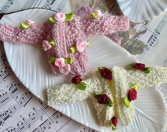 May include: Two miniature knitted cardigans with floral embellishments. One cardigan is pink with white and pink flowers, the other is white with green and red flowers.