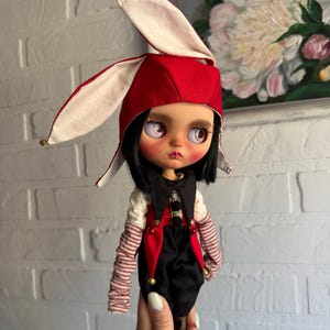 Pu&ograve; includere: Una bambola con capelli neri e occhi rosa indossa un cappello da giullare rosso e bianco con piccoli campanellini dorati. La bambola indossa un gilet nero e rosso sopra una camicia bianca con maniche lunghe a righe sottili rosse e bianche.