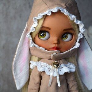 Puede incluir: Una mu&ntilde;eca con cabello rubio que lleva un atuendo marr&oacute;n con capucha y orejas de conejo blancas. La mu&ntilde;eca tiene ojos verdes y pecas.