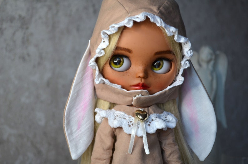 Puede incluir: Una mu&ntilde;eca con cabello rubio que lleva un atuendo marr&oacute;n con capucha y orejas de conejo blancas. La mu&ntilde;eca tiene ojos verdes y pecas.