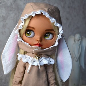 Puede incluir: Una mu&ntilde;eca con cabello rubio que lleva un vestido marr&oacute;n con capucha, ribete de encaje blanco y orejas de conejo. La mu&ntilde;eca tiene ojos marrones y pecas.