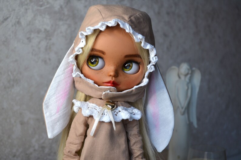 Puede incluir: Una mu&ntilde;eca con cabello rubio que lleva un vestido marr&oacute;n con capucha, ribete de encaje blanco y orejas de conejo. La mu&ntilde;eca tiene ojos marrones y pecas.